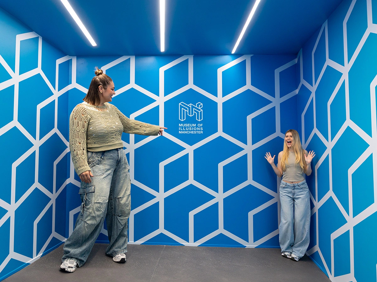Two women are standing in the corners of the Ames Room at Museum of Illusions Manchester. The woman in the left corner looks disproportionately tall compared to the woman in the right corner.