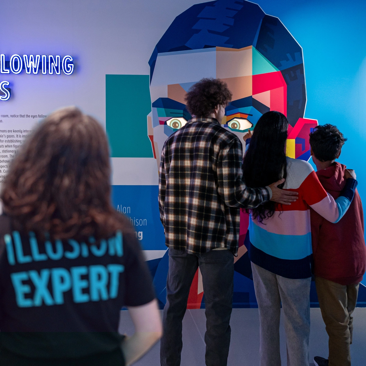 A family of three and an illusion expert at Museum of Illusions Manchester are looking at the Following Eyes exhibit. The exhibit represents Alan Turing.