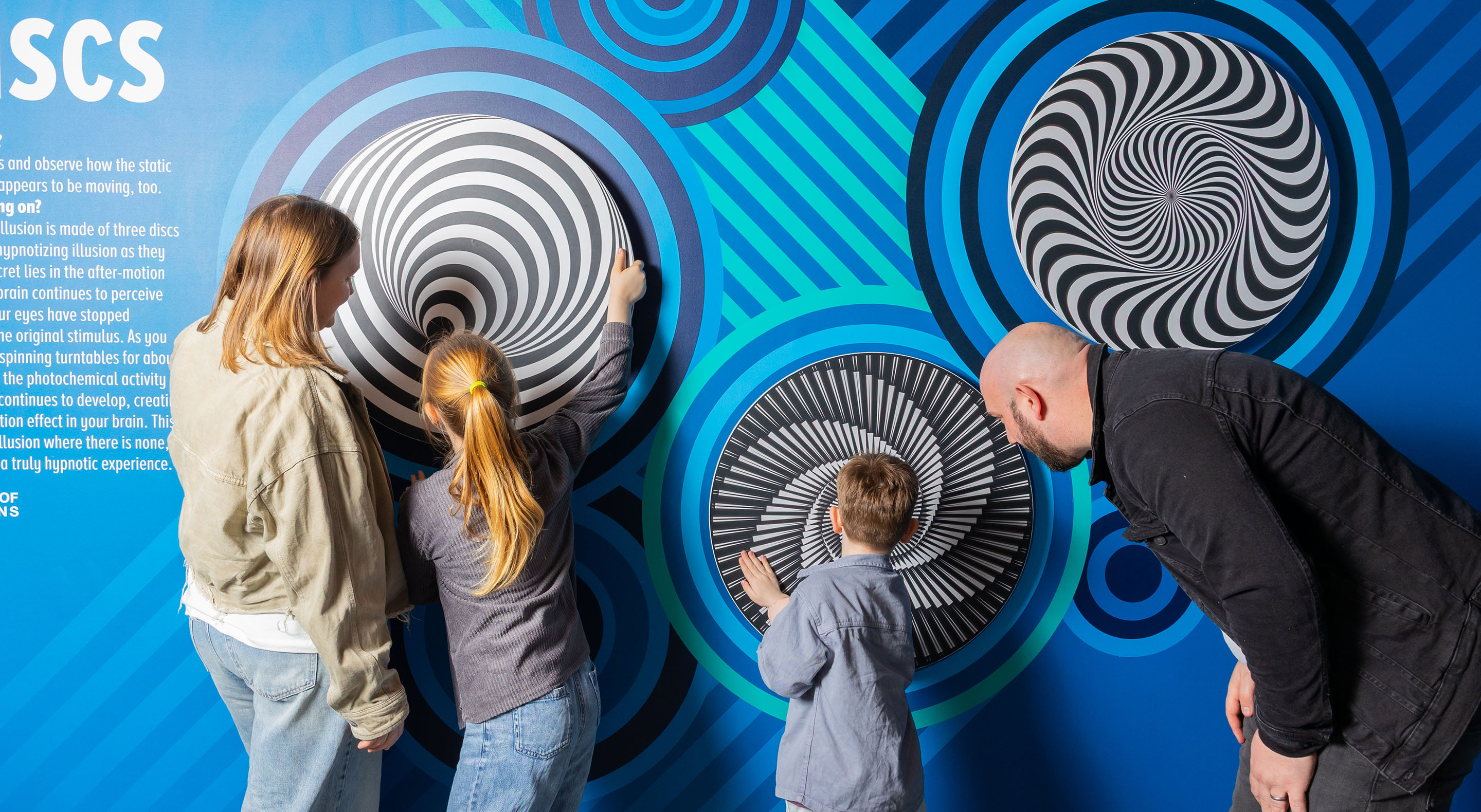 Parents with children are interacting with rotating illusion wheels on the wall.